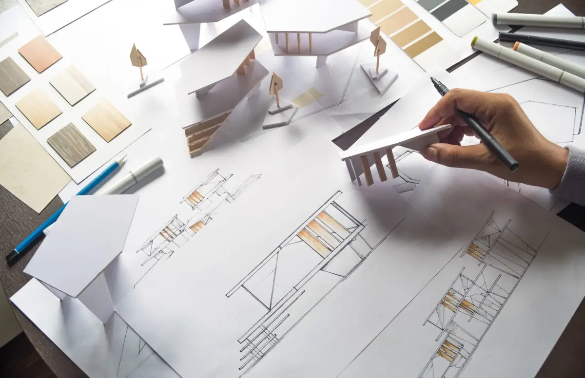 Ręka osoby rysującej model architektoniczny z planami i próbkami materiałów na biurku.