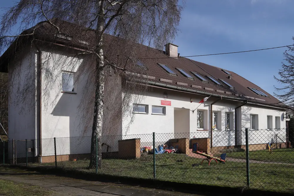 Widok na budynek przedszkola z białymi ścianami i brązowym dachem; po lewej stronie widać zabawki na placu zabaw za metalowym ogrodzeniem.